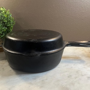 May include: A black cast iron double cooker set consisting of a deep round pot and a matching shallow skillet lid. The pieces feature integrated side handles and a long, flat handle on the skillet lid, with a smooth, matte finish throughout.