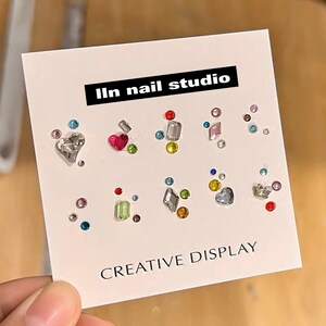 May include: A card with the text "lln nail studio" and "CREATIVE DISPLAY" features an assortment of colorful nail art rhinestones. The rhinestones include various shapes like hearts, rectangles, and circles, in shades of red, pink, silver, green, and blue.