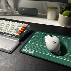 May include: A white wireless computer mouse with a black scroll wheel and logo sits on a rectangular green cutting mat mousepad featuring white grid lines, a barcode, and text that reads Industrial Inc. Cutting Mats. A clear mechanical keyboard is nearby.