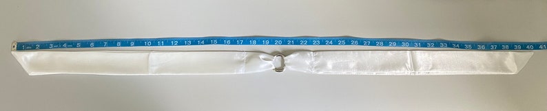 Puede incluir: Una faja de sat&eacute;n blanco con una hebilla plateada. La faja mide 104 cm de largo.