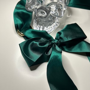 Puede incluir: Una cinta de sat&eacute;n verde oscuro atada en un lazo con un dije de calavera de cristal.