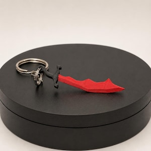 Op de afbeelding: Een rood-zwarte sleutelhanger in de vorm van een dolk. Het lemmet is rood met een gekartelde rand en het handvat is zwart. De sleutelring is zilverkleurig. Het item wordt gepresenteerd op een zwart, cirkelvormig oppervlak.