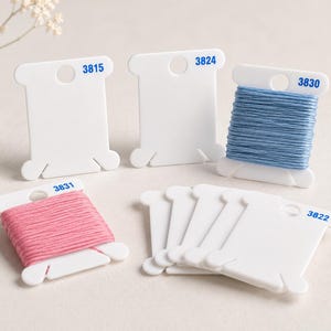 Op de afbeelding: Een verzameling witte plastic borduurgarenklosjes, sommige leeg en sommige omwikkeld met garen. Twee klosjes hebben blauwe gedrukte nummers 3831 en 3830 met roze en blauw garen, terwijl andere de nummers 3815, 3824 en 3822 tonen.