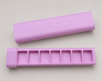 May include: A lavender pill organizer with a matching lid. The organizer is rectangular with seven compartments labeled for each day of the week. The lid is embossed with the name "CAROLA".