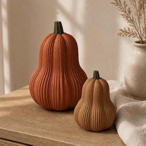 Op de afbeelding: Twee geribbelde pompoenvormige decoratieve objecten met donkergroene steeltjes. De grotere pompoen is feloranje, terwijl de kleinere een gedempte bruine kleur heeft. Beide hebben een getextureerd oppervlak met verticale ribbels.