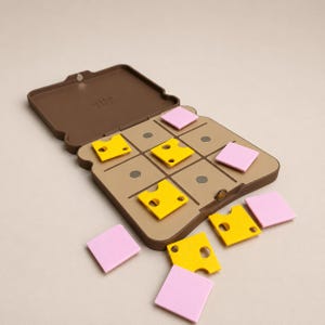 May include: A brown tic-tac-toe game set shaped like a sandwich, with yellow cheese pieces and pink bread pieces. The game is open, with some pieces scattered around. The game's lid is open, revealing the playing grid.