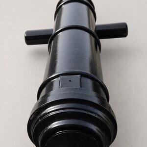 May include: A black, miniature cannon with a glossy finish. The cannon features a rounded base, a cylindrical barrel, and two small handles. The design is reminiscent of historical weaponry.