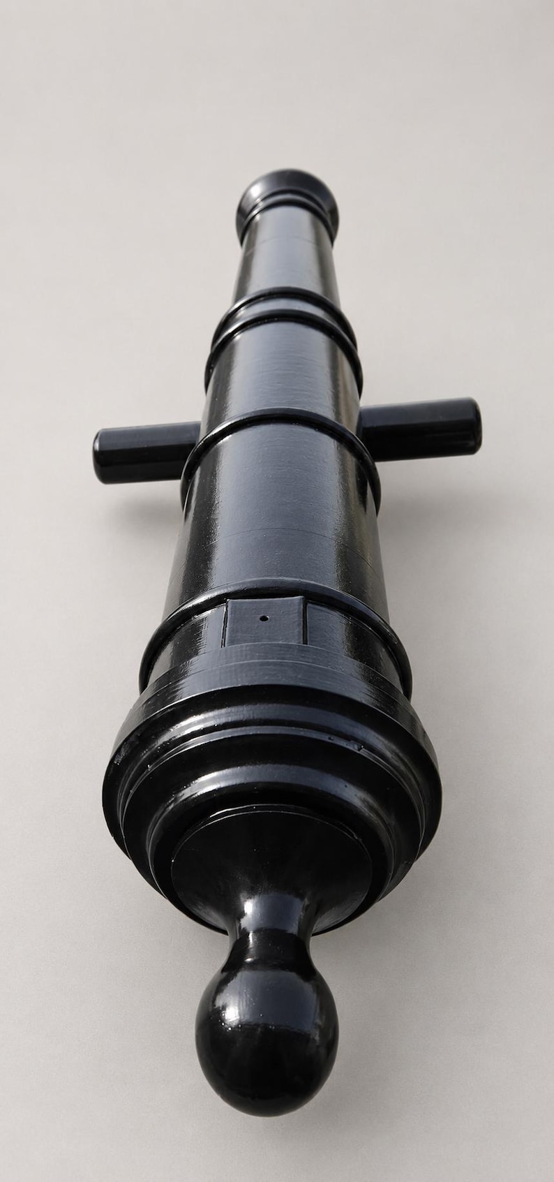 May include: A black, miniature cannon with a glossy finish. The cannon features a rounded base, a cylindrical barrel, and two small handles. The design is reminiscent of historical weaponry.