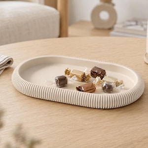 Pode incluir: Uma bandeja de formato oval na cor branco-sujo fosco, apresentando uma borda externa canelada e texturizada. A bandeja é rasa, com uma superfície interna lisa e plana, contendo vários pequenos bombons de chocolate embalados individualmente.