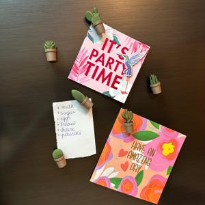 Könnte beinhalten: Kaktusförmige Magnete halten Notizen und Karten. Eine Karte zeigt "IT'S PARTY TIME" und eine andere sagt "HAVE AN AMAZING DAY". Eine Einkaufsliste wird ebenfalls von einem Magneten gehalten.