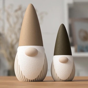 Op de afbeelding: Twee kabouterbeeldjes met gestructureerde, kegelvormige hoeden in beige en donker olijfgroen. Elke kabouter heeft een gladde, ronde neus, een gestructureerde witte baard en een basis met verticale ribbels in een lichte crèmekleur.