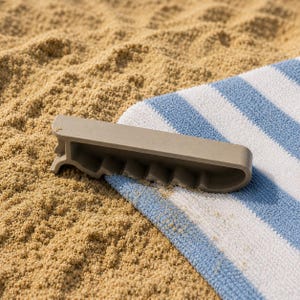 Op de afbeelding: Een lichtgrijze strandhanddoekclip ligt op een blauw-wit gestreepte handdoek, gedeeltelijk begraven in het zand. De clip heeft een gebogen ontwerp met meerdere sleuven om de handdoek vast te zetten. De scène roept een ontspannen dag op het strand op.