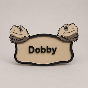 Peut inclure: Une étiquette nominative beige avec le nom "Dobby" en lettres noires. L'étiquette est encadrée en noir et comporte deux têtes de dragons barbus de dessin animé en haut. Les dragons barbus sont de couleur beige et marron.