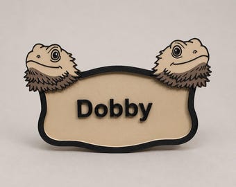 May include: A beige name tag with the name "Dobby" in black lettering. The tag is framed in black and features two cartoon bearded dragon heads at the top. The bearded dragons are tan and brown.