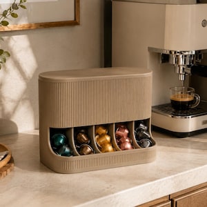 Op de afbeelding: Een beige, rechthoekige koffiecup-organizer met verticale ribbels. De unit heeft vijf open vakken aan de voorzijde, elk met een metalen koffiecup in verschillende kleuren, waaronder blauw, goud en roze.