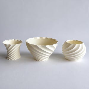 Op de afbeelding: Drie crèmekleurige keramische vazen met een spiraaltextuur. De vazen hebben een uniek, geribbeld ontwerp, met een cilindrische, komvormige en bolvormige vorm. De binnenkant van elke vaas is een lichtere tint crème.