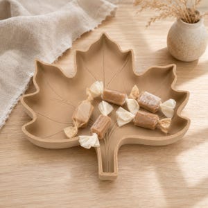 Op de afbeelding: Een mat beige, bladvormig dienblad met reliëf nerven. Het dienblad bevat verschillende rechthoekige, lichtbruine snoepjes verpakt in doorschijnend, gebroken wit papier met gedraaide uiteinden, gerangschikt in het midden.