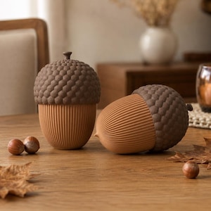 Op de afbeelding: Twee eikelvormige houders met gestructureerde bruine doppen en geribbelde beige onderkanten. De ene houder staat rechtop terwijl de andere op zijn kant ligt. De objecten hebben een gedetailleerd, herhalend patroon op de dop en verticale lijnen.