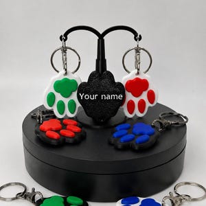 Op de afbeelding: Sleutelhangers in de vorm van een pootafdruk, met verschillende kleuren, waaronder groen, rood en blauw. De sleutelhangers zijn bevestigd aan zilveren sleutelringen. De tekst "Your name" is zichtbaar op een zwarte standaard.