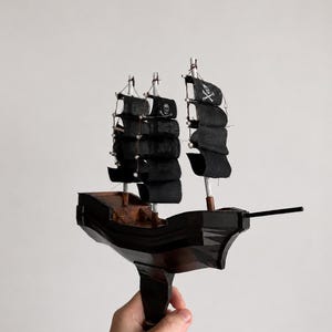 May include: A miniature wooden pirate ship model with black sails and a skull and crossbones flag. The ship has three masts and a dark brown hull. The model is held against a plain white background.