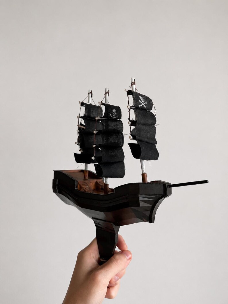 May include: A miniature wooden pirate ship model with black sails and a skull and crossbones flag. The ship has three masts and a dark brown hull. The model is held against a plain white background.