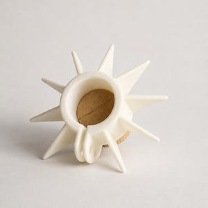 May include: A white, starburst-shaped bottle stopper with a cork base. The stopper features multiple pointed projections radiating outwards from a central ring. The design is modern and decorative, suitable for wine bottles.