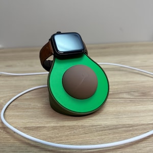 Op de afbeelding: Een groene avocado-vormige oplaadstandaard met een bruine pit, met een zwarte smartwatch met een bruine leren band. Een witte oplaadkabel is aangesloten op de standaard. De standaard staat op een houten oppervlak.