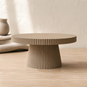 May include: A beige cake stand featuring a flat circular top and a tapered pedestal base, both decorated with a vertical ribbed texture. The object has a matte finish and a consistent neutral colour throughout its entire structure.