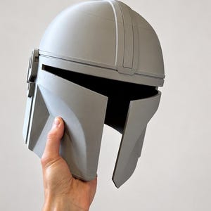 May include: A gray Mandalorian helmet, a replica of the iconic headgear. The helmet features a rounded top, a visor, and angular cheek plates. The helmet is held up against a plain background.