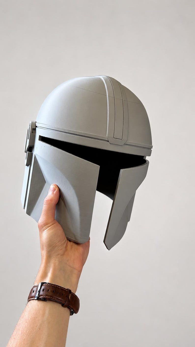 May include: A gray Mandalorian helmet, a replica of the iconic headgear. The helmet features a rounded top, a visor, and angular cheek plates. The helmet is held up against a plain background.