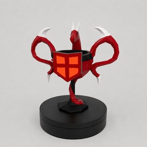 May include: A red and black geometric trophy with white accents and a shield design. The trophy is mounted on a black circular base. The shield has a red cross on an orange background.