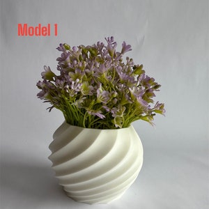 Op de afbeelding: Witte keramische vaas met een draaiend, gestructureerd ontwerp. De vaas is gevuld met een boeket kunstmatige paarse en groene bloemen. De tekst "Model 1" is zichtbaar in de linkerbovenhoek van de afbeelding.