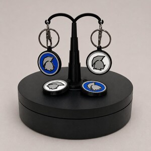 May include: Keychains with a circular design featuring a gray helmet on a white or blue background. The keychains are attached to silver key rings. Two keychains are hanging, and two are resting on a black platform.