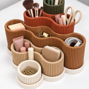 Pode incluir: Um organizador de secretária de vários níveis com formato ondulado e textura canelada em tons de creme, bege, castanho claro, terracota e verde azeitona. Contém vários compartimentos com pincéis de maquilhagem, canetas, tesouras e cabos.