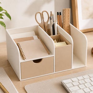 May include: A white and beige desk organiser featuring a small drawer, multiple vertical compartments for stationery, and open slots for notebooks. The unit holds scissors, pens, and a wooden ruler, providing a structured storage solution for office supplies.