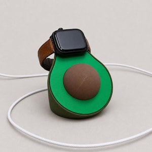 Op de afbeelding: Een groene avocado-vormige oplaadstandaard met een bruine pit, met een zwarte smartwatch met een bruine leren band. Een witte oplaadkabel is aangesloten. De standaard is ontworpen voor smartwatches.
