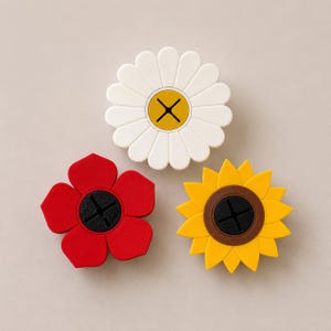Op de afbeelding: Drie bloemvormige siliconen onderzetters. Eén is wit met een gele kern, een andere is rood en de derde is geel met een bruine kern. Elke onderzetter heeft een zwart kruisvormig ontwerp in het midden.