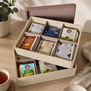 Op de afbeelding: Een beige theedoos met een deksel, verdeeld in compartimenten gevuld met diverse theezakjes. De theezakjes hebben kleurrijke verpakkingen met teksten als "Zen Chai" en "Rooibos". Een kopje thee en een schoteltje zijn zichtbaar.