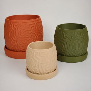 Op de afbeelding: Drie cilindrische plantenpotten met bijpassende schotels, elk voorzien van een verhoogd, doorlopend, hersenachtig doolhofpatroon aan de buitenkant. De set bevat een grote terracotta oranje pot, een middelgrote olijfgroene pot en een kleine beige pot.