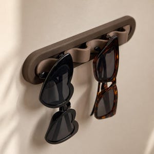 May include: A dark brown wall-mounted organizer rack with a wavy, multi-slot design for hanging eyewear. Two pairs of sunglasses are suspended from the rack: one pair with black heart-shaped frames and one pair with brown tortoiseshell frames.