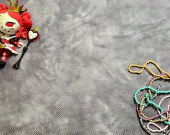 May include: A stylized red-haired queen figurine wearing a gold crown and a red and white dress, holding a heart-topped scepter, positioned next to a cluster of multi-colored beaded necklaces on a textured gray fabric background.