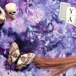 May include: An overhead shot features a doll head, a skull, a butterfly with a skull design, and playing cards with the letter "Y" on a purple and white tie-dye background. A strand of brown hair is also present.
