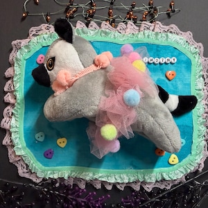 May include: A plush raccoon toy with a black and white striped tail, wearing a pink tulle skirt adorned with colourful pom-poms and a pink trim collar. The toy is placed on a rectangular turquoise fabric mat with lace edges and the name TIMOTHY.