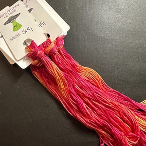 Puede incluir: Haces de hilo de bordar en vibrantes tonos de rosa, naranja y amarillo. El hilo está anudado y sujeto a tarjetas blancas. Las tarjetas tienen el texto "Aliens Among Us Floss" y un gráfico de un OVNI.