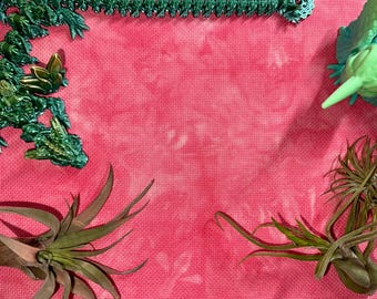 May include: A piece of pink fabric with a mottled tie-dye pattern, featuring a metallic green dragon figure along the top and left edges, a light green creature on the top right, and two spiky air plants positioned at the bottom corners.