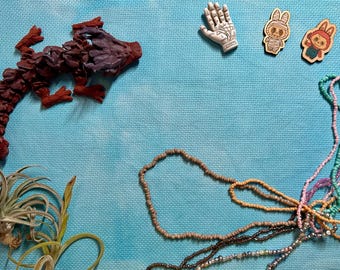 May include: A dark red dragon figurine, a white palmistry hand charm, two small wooden character pins, a green air plant, and several strands of colorful beaded necklaces arranged on a textured light blue surface.