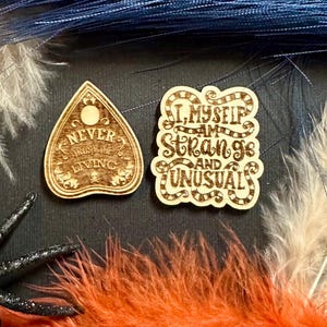 May include: Two wooden, laser-cut,  ouija board-shaped ornaments. One says "Never trust the living" and the other says "I, myself, am strange and unusual."