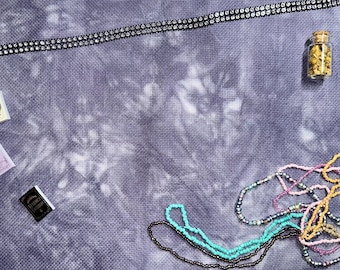 May include: An overhead shot features various craft supplies on a textured, tie-dye fabric. A sparkling rhinestone trim, miniature books, a small glass bottle filled with gold-colored beads, and strands of colorful beads are scattered across the surface.