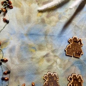 May include: Three laser-cut wooden haunted house shapes with ghost details, arranged on a mottled blue and yellow fabric background. A string of small orange lights with dark bases curves along the left side of the fabric surface.