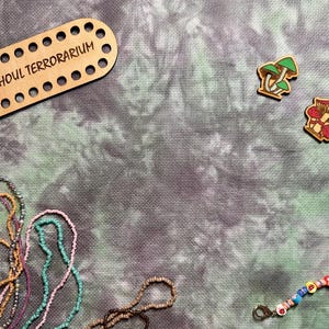 May include: Wooden craft supplies and beads on a textured background. Includes a wooden oval with the text "TOADGHOUL TERRARIUM", mushroom-shaped wooden embellishments, and strands of colorful beads.